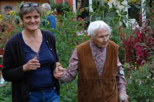 Foto; zwei Frauen spazieren im Garten, die jüngere Frau reicht der älteren Frau die Hand als Stütze beim Laufen