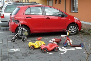 Foto; Parkplatz, im Hintergrund ein roter Kleinwagen, im Vordergrund ein Rollator, ein Dreirad, zwei Bobby-Cars und ein Kinderroller